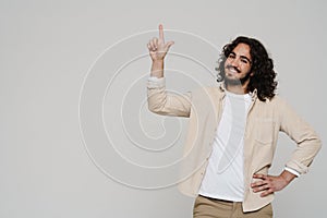 Smiling hispanic man pointing finger up at copy space while standing isolated over white wall