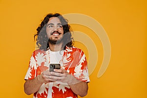 Smiling hispanic man using smartphone and looking away isolated over yellow background