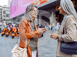 Smiling mature women and Asian lady with modern mobile phones talk on city street