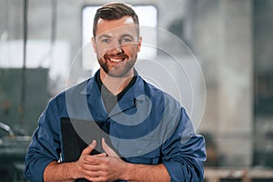 Smiling and holding tablet in hands. Factory worker in blue uniform is indoors