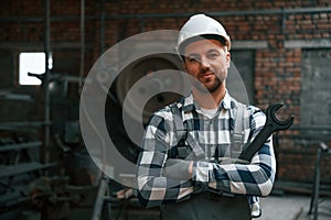 Smiling and holding wrench. Factory male worker in uniform is indoors