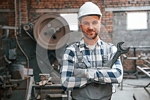 Smiling and holding wrench. Factory male worker in uniform is indoors