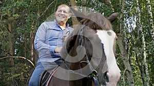 Smiling horseman sitting astride horse on green trees background on farm ranch. Happy stableman sitting on horseback