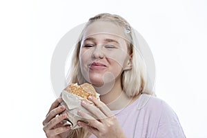 Smiling hungry teenage girl takes a bite of hamburger. Delicious popular fast food. A cute blonde with freckles in a pink jacket.