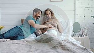 Smiling husband stroking pregnant belly of wife. Pregnant couple huggins in bed