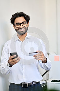 Smiling indian business man using phone holding credit card making e payment.