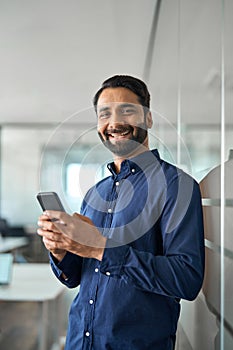 Smiling Indian businessman using mobile cell phone standing in office. Vertical