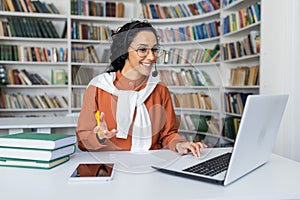 Smiling Indian businesswoman in headset sits at laptop in room, communicates with clients online, works in call center