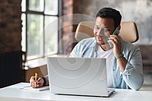 Smiling indian freelancer guy is talking on smartphone and taking notes