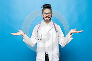 Smiling indian male doctor holding palms upwards as copyspace isolated on blue background