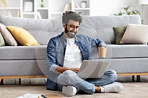 Smiling Indian man using laptop while sitting cross-legged on floor at home