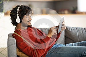 Smiling Indian Man Wearing Wireless Headphones Resting With Digital Tablet At Home