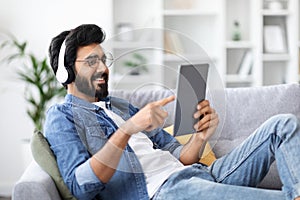 Smiling Indian Man Wearing Wireless Headphones Resting With Digital Tablet At Home
