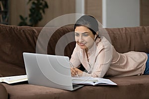 Smiling Indian woman in earphones studying online, lying on couch