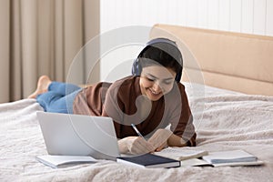 Smiling Indian woman in headphones taking notes, lying on bed