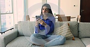Smiling Indian woman relax sit on couch with cellphone