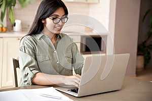 Smiling indian young woman typing on laptop computer working at home office.