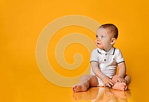 Smiling infant baby boy toddler is sitting on the floor looking aside at free copy space on yellow background