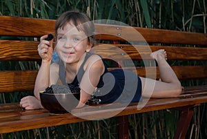 Smiling ittle girl lies on a wooden bench with a berry in her hand.