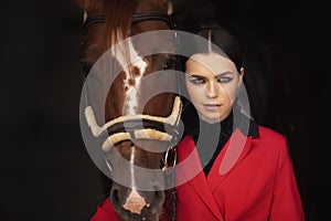 Smiling jockey woman standing with brown horse in stable, black background