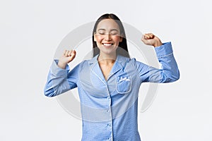 Smiling joyful asian girl in pajamas standing with closed eyes and delighted grin, stratching after good sleep, waking