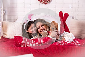 Smiling joyful mom with daughter in Christmas ears with soft toy teddy bear in bed with illuminations New Year, 2020, Christmas