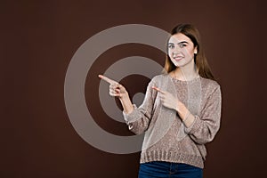 Smiling joyful woman in loose sweater posing against brown studio wall pointing at copy space for advertisment or photo