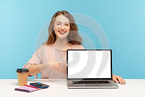 Smiling joyful woman, office employee, sitting at workplace and pointing empty laptop screen, mock up display