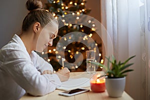 Smiling joyful woman in white shirt sitting at table and writing Christmas bucket list or tasks she needs to do in preparation for