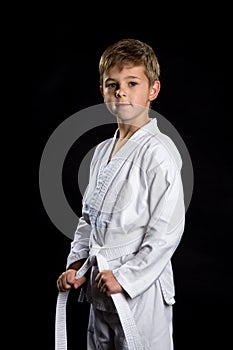 Smiling karate kid in brand new kimono, posing on the black background. Karate fighter holding his belt