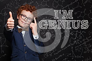 Smiling kid boy with red hair thinking, pointing at his head and standing against blackboard with science formulas
