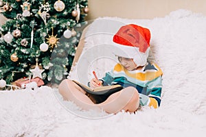 Smiling Kid boy in red Santa hat sit on couch and writing the letter to dear Santa at home. Child wish list. Dreams of a