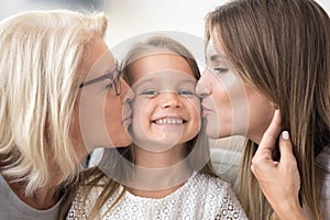 Smiling kid girl enjoys mom and grandma kissing on cheeks