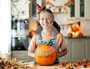 Smiling kid girl in hat showing pumpkin with inscription Happy halloween during Halloween celebration