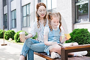 smiling kid with juice reading book with mother near by while resting on bench together