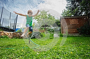 Smiling kid jump over a sprinkler on summer lawn in backyard