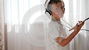 Smiling kid with phone into hands listening to music across headset and dancing in room