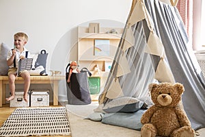 Smiling kid sitting on bench with notebook, real photo of natural playroom interior with scandinavian tent and teddy bear