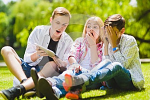 Smiling kids having fun and look to tablet at grass. Children playing outdoors in summer. teenagers communicate outdoor