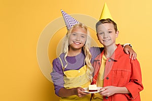 Smiling kids with party caps holding plate with birthday cake and hugging