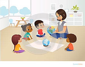 Smiling kindergarten teacher shows globe to children sitting in circle during geography lesson. Preschool activities