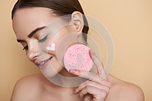 Smiling lady with facial cream on cheek showing round pink sponge