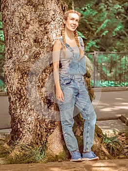 Smiling lady with pigtails in denim overalls stands leaning on the trunk of a massive tree in the park on a summer day