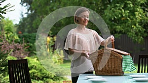 Smiling lady puts hamper on table, family dinner outdoors, picnic preparation