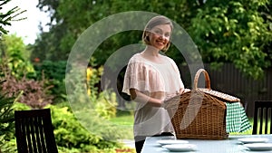 Smiling lady puts hamper on table, family dinner outdoors, picnic preparation