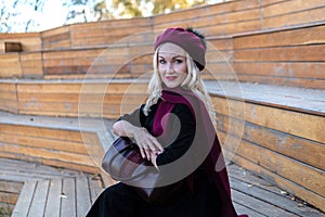 A smiling lady sits on a summer theater bench, wooden in a burgundy coat and biret, an adult with a beautiful smile, in