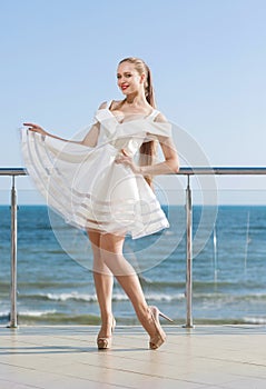 A smiling lady on a terrace. A happy female with a long ponytail on a sunny sky background. girl in a white dress