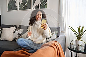 Smiling latin girl sitting on couch using cell phone in living room at home.