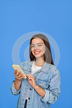 Happy Latin teen girl wearing denim using phone on blue background. Vertical.