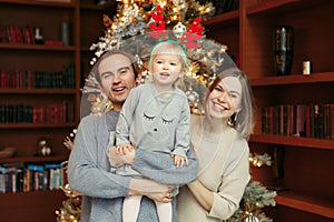 Smiling laughing Caucasian mother and father with baby girl playing together by decorated Christmas tree. Happy family celebrating